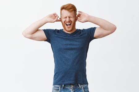 Displeased redhead guy feeling pain in head cause of noisy annoying sound, covering ears with fingers, shouting from painful feeling, closing eyes and yelling with opened mouth over grey wallの写真素材