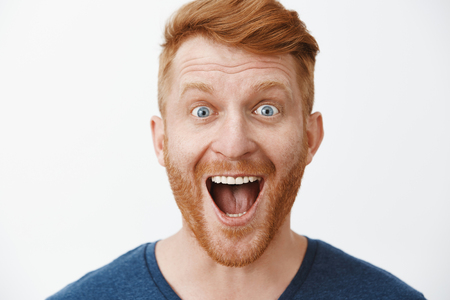 Close-up shot of amazed and joyful adult redhead male model with beard smiling broadly with dropped jaw and impressed expression, standing pleased and thrilled from positive event over grey wallの写真素材