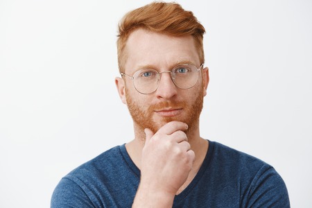 Headshot of creative and smart handsome redhead guy with bristle in glasses and blue t-shirt, rubbing beard on chin and gazing with smirk at camera, having great plan or idea over gray backgroundの写真素材