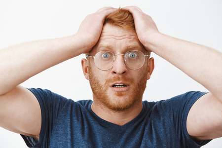 Headshot of worried attractive redhead businessman in glasses and blue t-shirt holding hands on hair and frowning, standing in stupor at shocking scene, feeling anxious and desperateの写真素材