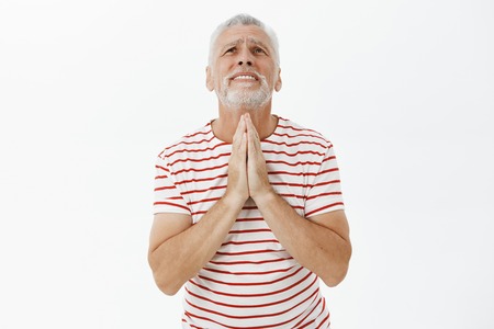 Hopeful old man in need and trouble wearing striped t-shirt with grey beard holding hands in pray looking up in sky praying god with concerned and faithful expression over gray backgroundの写真素材