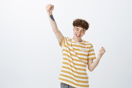 Yes I am winner. Portrait of charming delighted and excited young curly-haired male student with tattoo raising up fist in triumph and victory smiling achieving success grinning from delightの写真素材