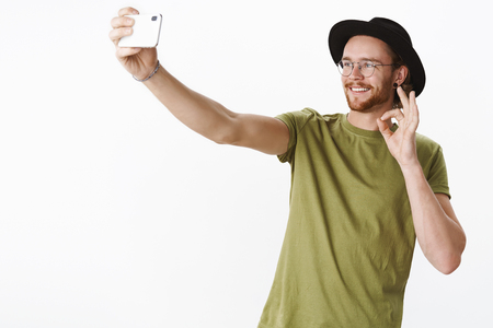 Studio shot of cute male traveler on vacation taking selfies on smartphone extanding hand with mobile phone and smiling at screen showing okay gesture as being glad and satisfied with tripの写真素材