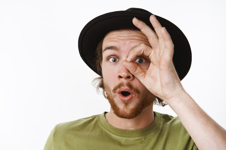 Wow look at this prices. Impressed and surprised charismatic handsome young male with beard and pierced nose folding lips curiously as shown ok gesture over eye peeking through finger holeの写真素材