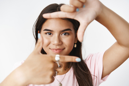 Close-up shot of imaginative and creative good-looking indian girl making frames with fingers and looking through it as creating idea or plan what paint or how shoot with camera over gray wallの写真素材