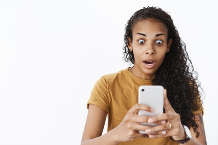 Waist-up shot of shocked astonished and speechless african american young woman with tattoo dropping jaw gasping amazed as popping eyes at smartphone screen seeing feedback from superstarの写真素材