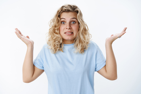 Portrait of awkward apologizing cute unaware and confused european blond female with blue eyes saying oops and grimacing as raising hands in clueless gesture over white wallの写真素材