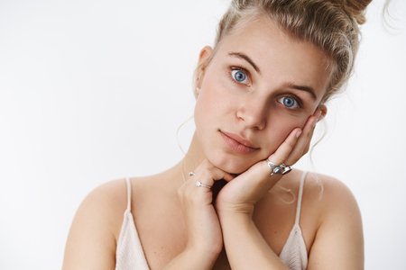 Flirty and tender blond girlfriend with hairbun and blue eyes tilting head leaning on palm gentle and cute making lovely gazes at camera as seducing being silly, expressing tenderness over white wallの写真素材