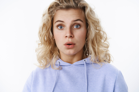 Close-up shot of wondered and surprised cute european female with blue eyes and blond curly hair raise eyebrows in amazement open mouth from astonishment, impressed over white wallの写真素材