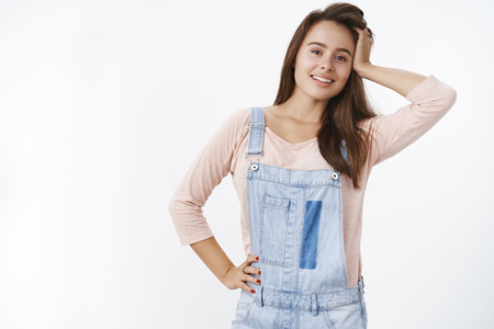Indoor shot of attractive brunette girlfriend in denim overalls playing with hair and holding hand on hip as smiling broadly at camera making cute gazes, flirting over gray backgroundの写真素材