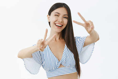 Waist-up shot of upbeat energized and emotive charming asian woman in cropped blouse showing peace or victory gestures winking happily and smiling at camera, having fun over white backgroundの写真素材
