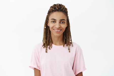 Calm and friendly good-looking young african american female with dreads in pink stylish t-shirt smiling assured and joyful feeling awesome and confident during casual talk over white backgroundの写真素材