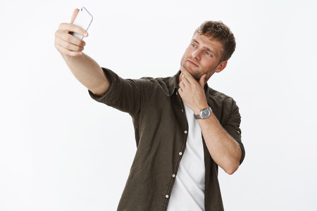 Handsome and masculine macho man making cool pose touching bristle on jaw and taking selfie with sexy smile at smartphone screen extending hand to make nice shot over gray backgroundの写真素材