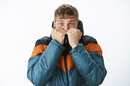 Scared good-looking european blond man with blue eyes getting warm pulling collar of puffer jacket on face exhaling air, standing outside on freezing cold weather over gray wallの写真素材