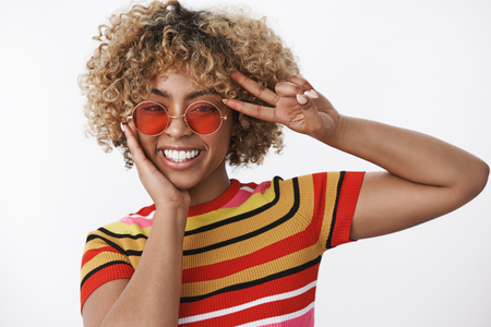 Hey peace guys. Carefree and joyful friendly-looking stylish african american female with red sunglasses and striped top showing victory or disco gesture near eye and smiling broadly over white wallの写真素材