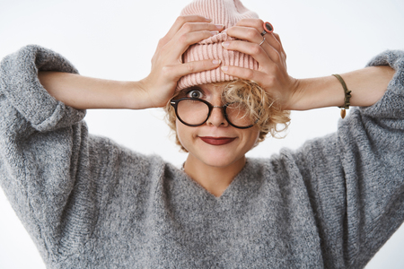 Close-up shot of modern and stylish beautiful trendy hipster girl pulling beanie on forehead smirking joyfully and looking at camera with happy carefree gaze holding hands on head over white wallの写真素材