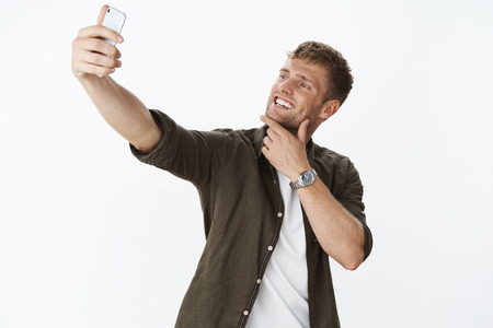Cool and stylish young handsome blond man touching chin and smiling broadly as extending hand to take selfie on smartphone making happy face, posing over gray background delightedの写真素材