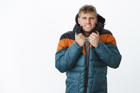 Jeez freezing cold. Portrait of intense trembling handsome european blond man clenching teeth wearing winter puffer jacket suffering from low temperature and bad weather posing over gray backgroundの写真素材