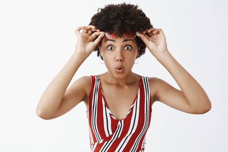 Impressed attractive African American girl with curly hairstyle, taking off sunglasses and staring curious and amazed at camera, folding lips from interestの写真素材