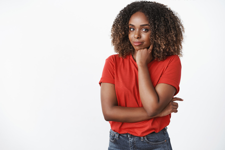 Indoor shot of creative and confident good-looking young african-american woman in red t-shirt leaning head on fist and smiling self-assured at camera having great plan how gain success in lifeの写真素材