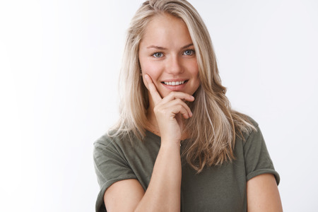 Close-up shot of powerful self-assured good-looking blond woman with tan and tattoo lean head on hand smiling intrigued and curious with cheeky gaze know how catch eye, flirting over white wallの写真素材