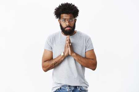 Sad and gloomy african american guy with pierced nose, tattoos wearing glasses pouting upset holding hands in pray over chest, frowning begging for mercy and help, asking favour or sorryの写真素材