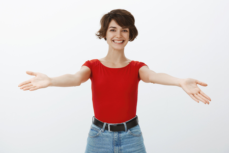 Girl welcomes dear guests spreads hands sideways smiling broadly greeting friend wanna give warm embraces cuddles happy see people comming party standing glad white background red t-shirtの写真素材