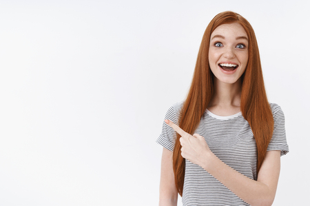 Amused surprised enthusiastic wondered redhead young cute ginger girl wearing striped t-shirt drop jaw gasping astonished widen eyes astonished pointing upper left corner thrilled, white backgroundの写真素材
