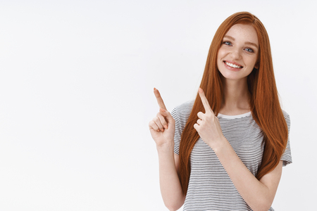Charming lovely kind redhead blue-eyed girl tilting head smiling broadly give advice recommend choose direction pointing upper left corner showing good choice, white backgroundの写真素材