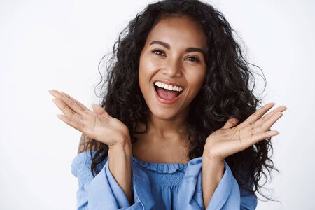Close-up emotive, tender and feminine surprised african-american curly-haired woman in blue blouse reacting to wonderful news, spread arms gladly, smiling broadly, stand white backgroundの写真素材
