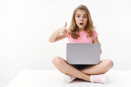 Shocked worried cute blond girl little daughter show mom scary video computer, sit crossed legs, hold laptop, pointing gadget screen, gasping scared and nervous, stare camera upset, white backgroundの写真素材