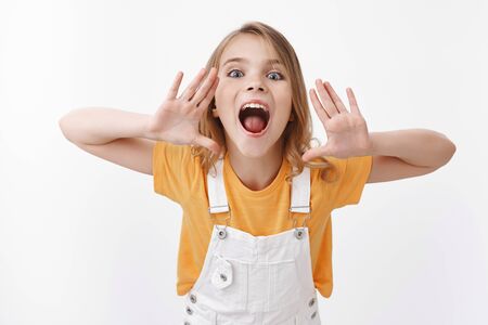 Cute pretty playful little girl, blond charming child searching someone, hold hands near opened mouth, shout loud at you camera, look forward, looking for friend, calling with yell, white backgroundの写真素材