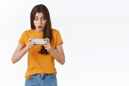 Shocked thrilled young brunette female playing intense battle game, holding smartphone horizontally tap mobile screen to win, gasping stare telephone display with popped eyes, white backgroundの写真素材