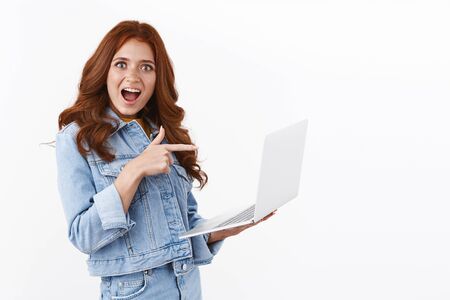 Amused enthusiastic redhead female in denim jacket, standing half-turned over white background, hold laptop pointing finger computer screen, checking out awesome offer internet, smiling astonishedの写真素材