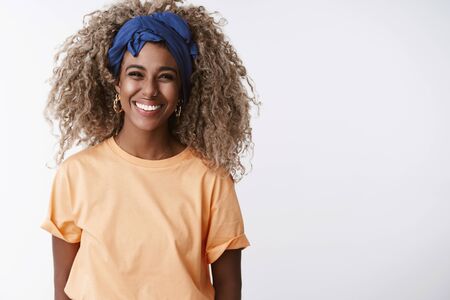 Waist-up shot friendly and sincere, attractive african-american blond girl with afro hairstyle, stylish headband and orange t-shirt, laughing and smiling joyful, have fun standing white backgroundの写真素材