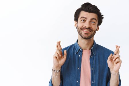 Close-up optimistic, good-looking brunette male with beard, cross fingers for good luck, smiling and gazing camera, anticipating something positive happen, believe dreams come true, white backgroundの写真素材