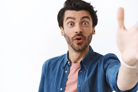 Charming, handsome bearded caucasian man with stylish haircut, holding camera with hand as taking seflie with surprised, joyful expression, record live video on smartphone, standing white backgroundの写真素材