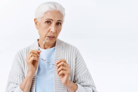Curious, thoughtful cute senior woman with grey combed neat haircut, take-off glasses, biting rim of...