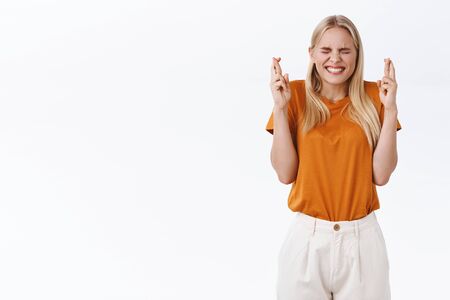 Hopeful cute, optimistic young blond girl in stylish t-shirt, pants, fingers crossed and smiling with closed eyes, making wish, awaiting dream come true, desire to gain achievement, white backgroundの写真素材