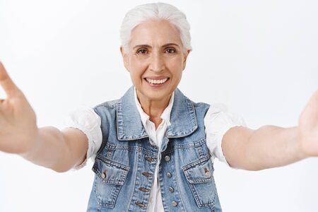 Friendly, tender and kind senior woman with grey combed hair, reaching hands forward, hold camera as...