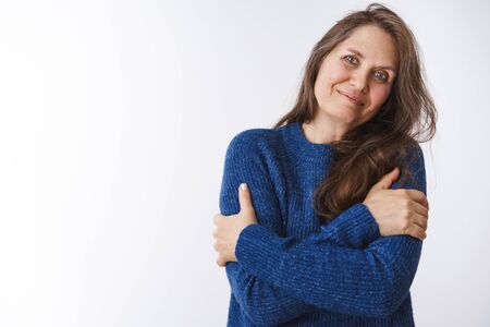 Granny waits family come see her on holidays hugging herself tight, tiltign head on shoulder and smiling cute, wearing blue warm sweater, feeling tender and gentle against white backgroundの写真素材
