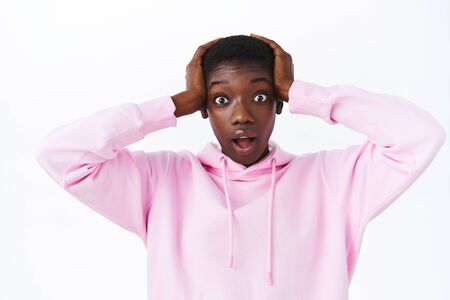 Mind-blowing news. Shocked african-american woman in pink hoodie grab head both arms and gasping, drop jaw and pop eyes looking at something extremely impressive, stand white backgroundの写真素材