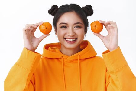 Close-up portrait of kawaii smiling young asian girl with two tangerines, giggle silly and looking...