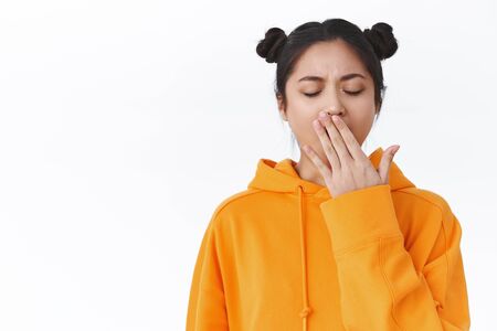 Tired cute female asian student study all night, didnt have much sleep, close eyes and yawning lazy, cover opened mouth with palm, standing white background in orange hoodie, need morning coffeeの写真素材