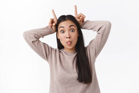 Playful gorgeous asian girl with long healthy dark hair, making horns on head and popping eyes...