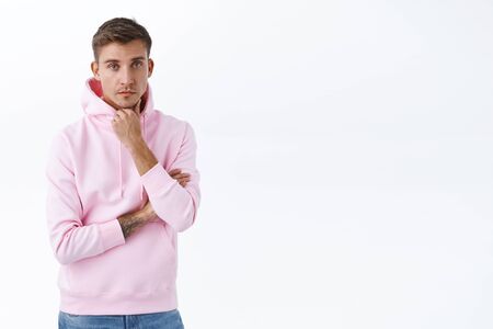 Serious-looking blond young man in pink hoodie, listening with skeptical look uninteresting, boring information, touch chin as thinking, making choice or plan thoughtful, stand white backgroundの写真素材