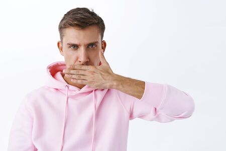 Young serious blond man cover his mouth with hand and look camera strict, fed up stay silent,...