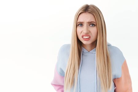 Close-up portrait of embarrassed blonde girl, stooping and clench fists, grimacing as staring something cringy, standing in awkward awful situation, want to escape, white backgroundの写真素材