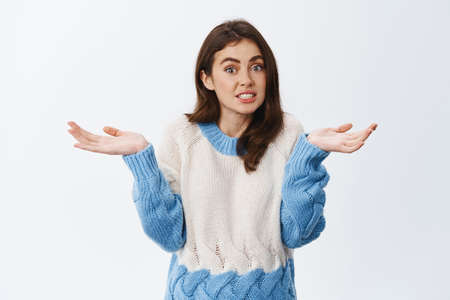 Silly young girl shrugging shoulders and spread hands sideways, being unaware and clueless, have no idea what happening, standing against white backgroundの写真素材