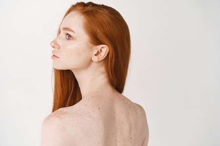 Portrait of beautiful young woman with long red healthy hair and pale skin, showing clean smooth back and looking left, standing on white backgroundの写真素材